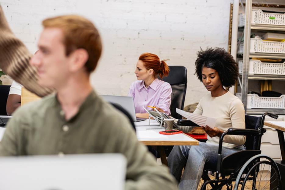 An office scene shows diverse employees at work. A woman in a wheelchair reviews paperwork while two other women work nearby at computers.